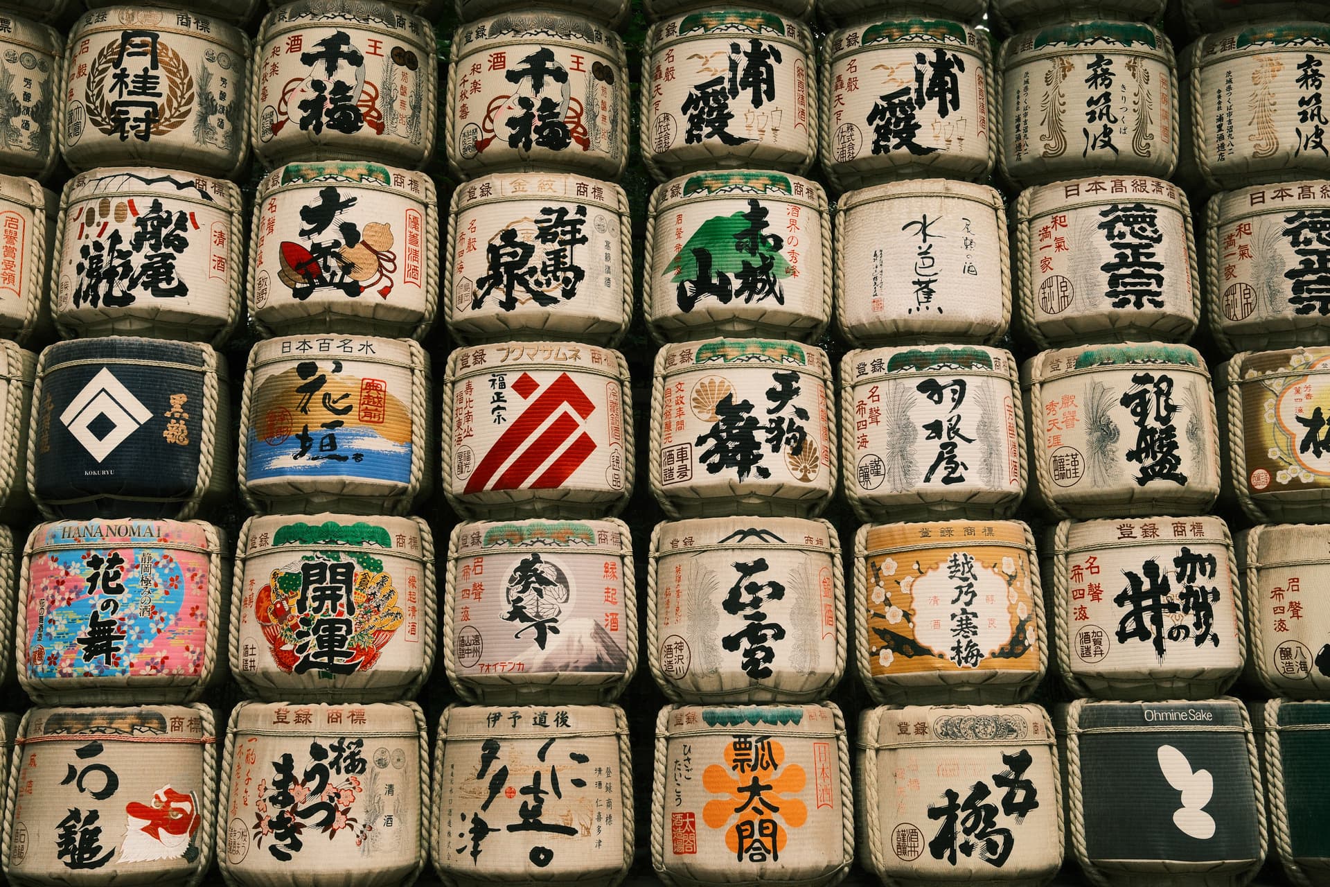 A grid of Japanese sake barrels (sake-daru) displays various brewery names and logos in traditional calligraphy, with white, beige, and occasional colorful backgrounds arranged in neat rows, representing different sake producers from across Japan.