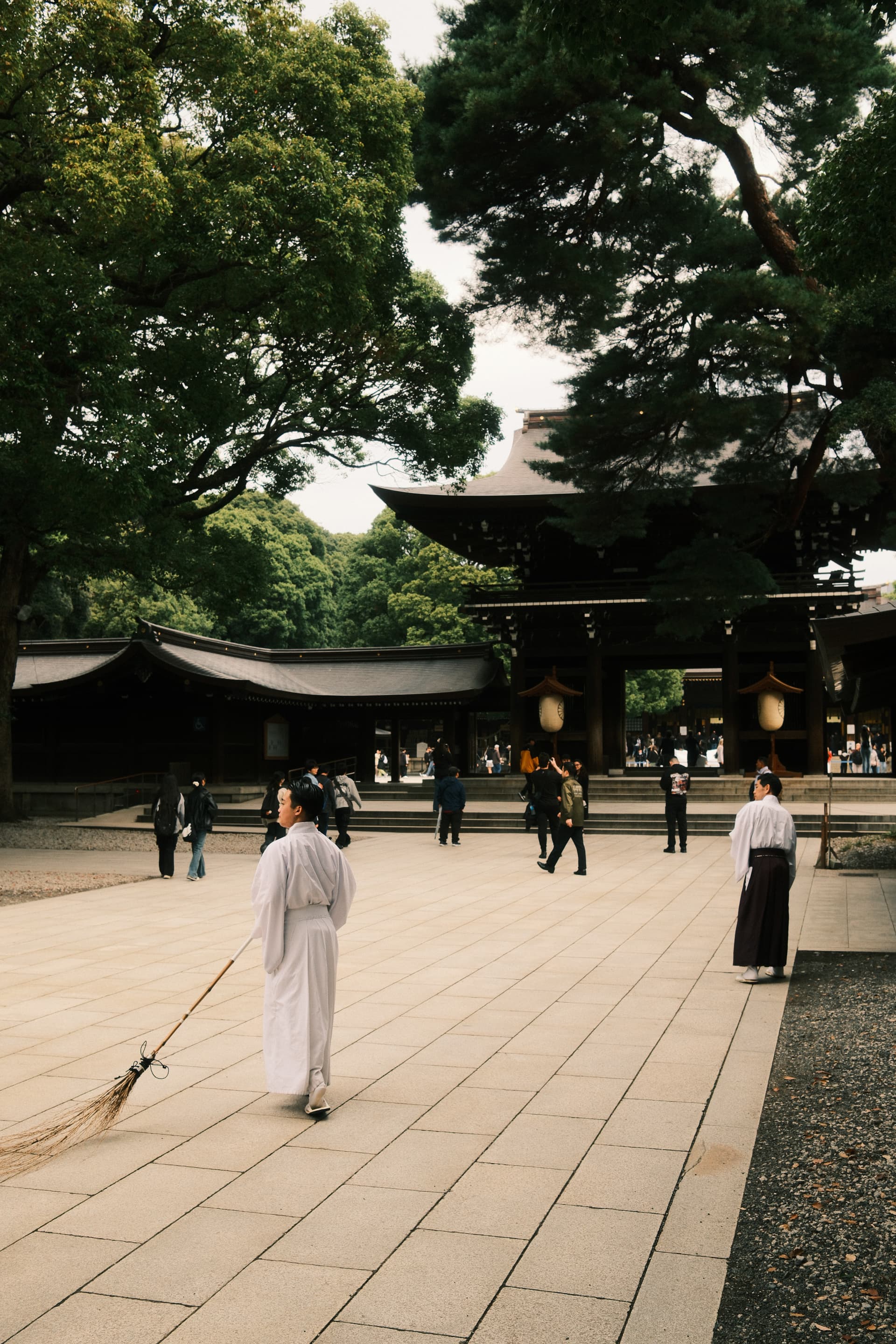A traditional Japanese shrine courtyard with stone-tiled ground, featuring a white-robed attendant sweeping near a wooden temple building, surrounded by lush green trees and several visitors walking quietly through the peaceful space.