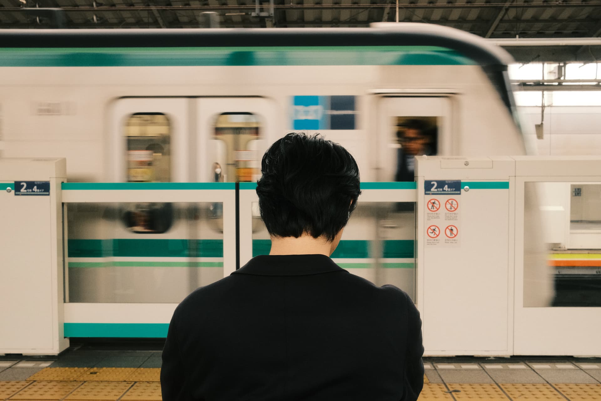 A person wearing black stands facing a blurred green and white metro train at a Tokyo subway station, with platform safety barriers and signage visible.