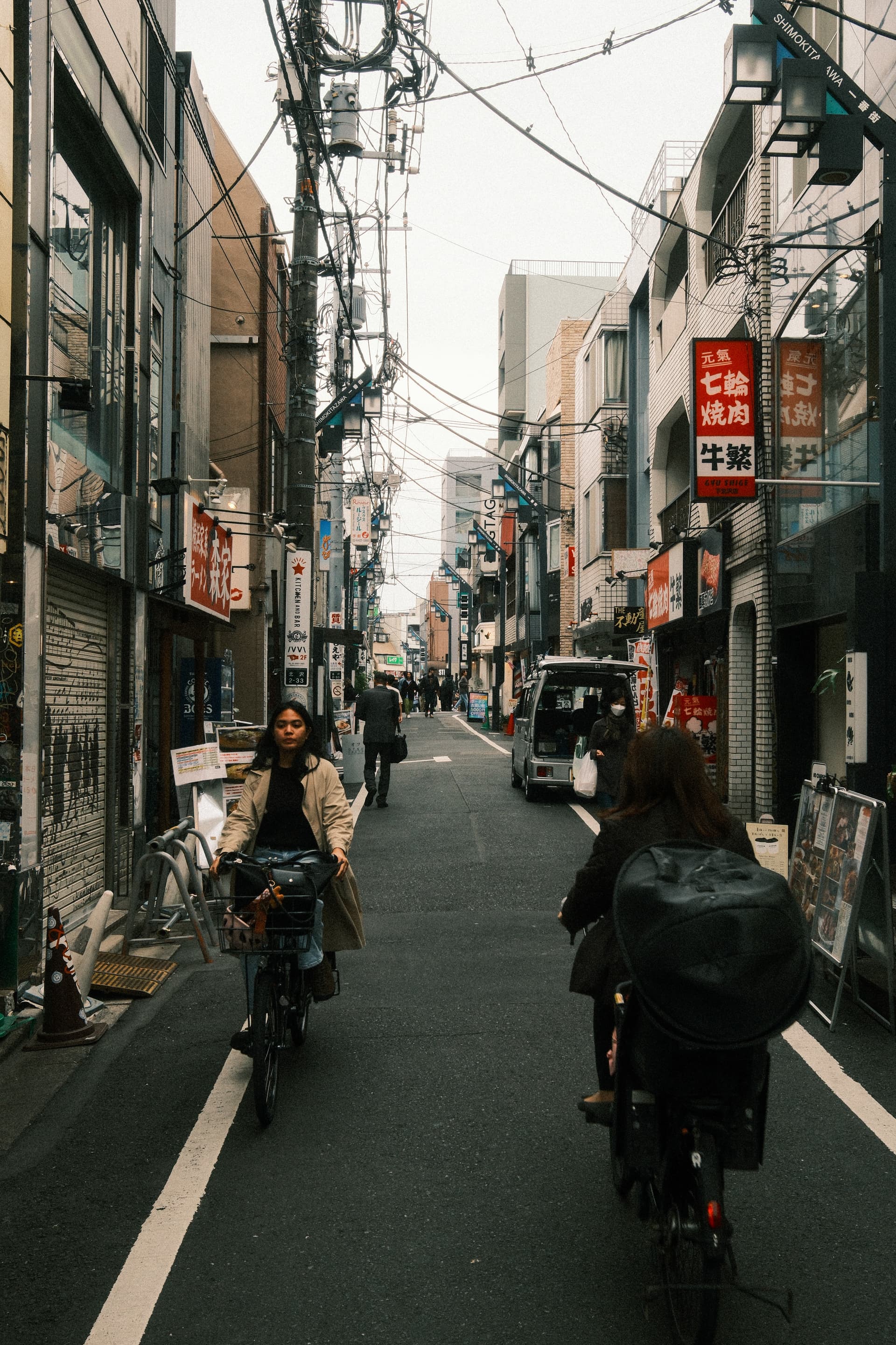 A narrow urban street in Tokyo lined with electrical wires, bicyclists, and red Japanese signage, capturing the dense, layered texture of a typical residential neighborhood with buildings and utility poles crowding the streetscape.