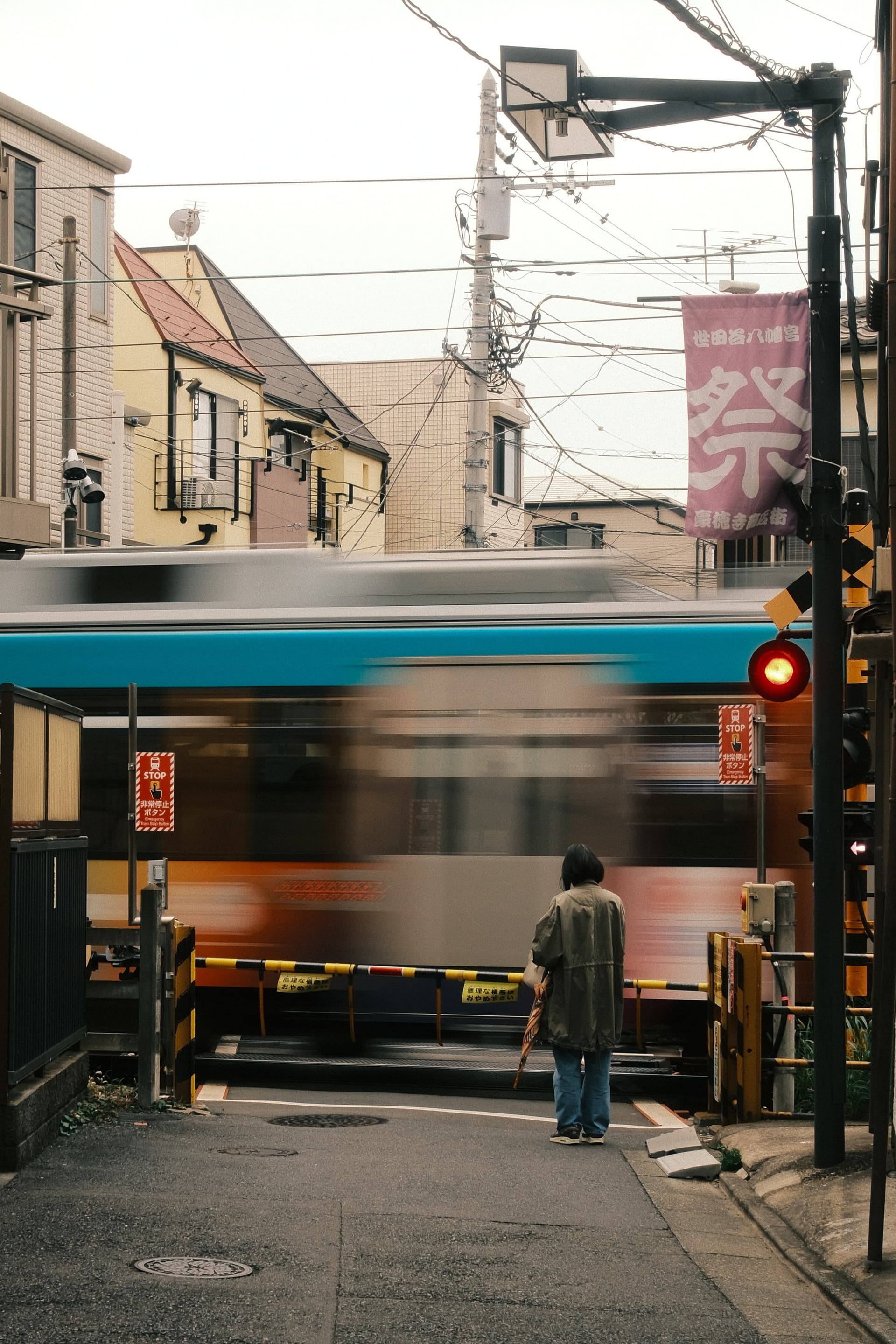 A blurred teal train passes through a crowded urban train crossing in Tokyo, with power lines overhead and a person standing near the gates, waiting against a backdrop of residential buildings and a pink festival banner.