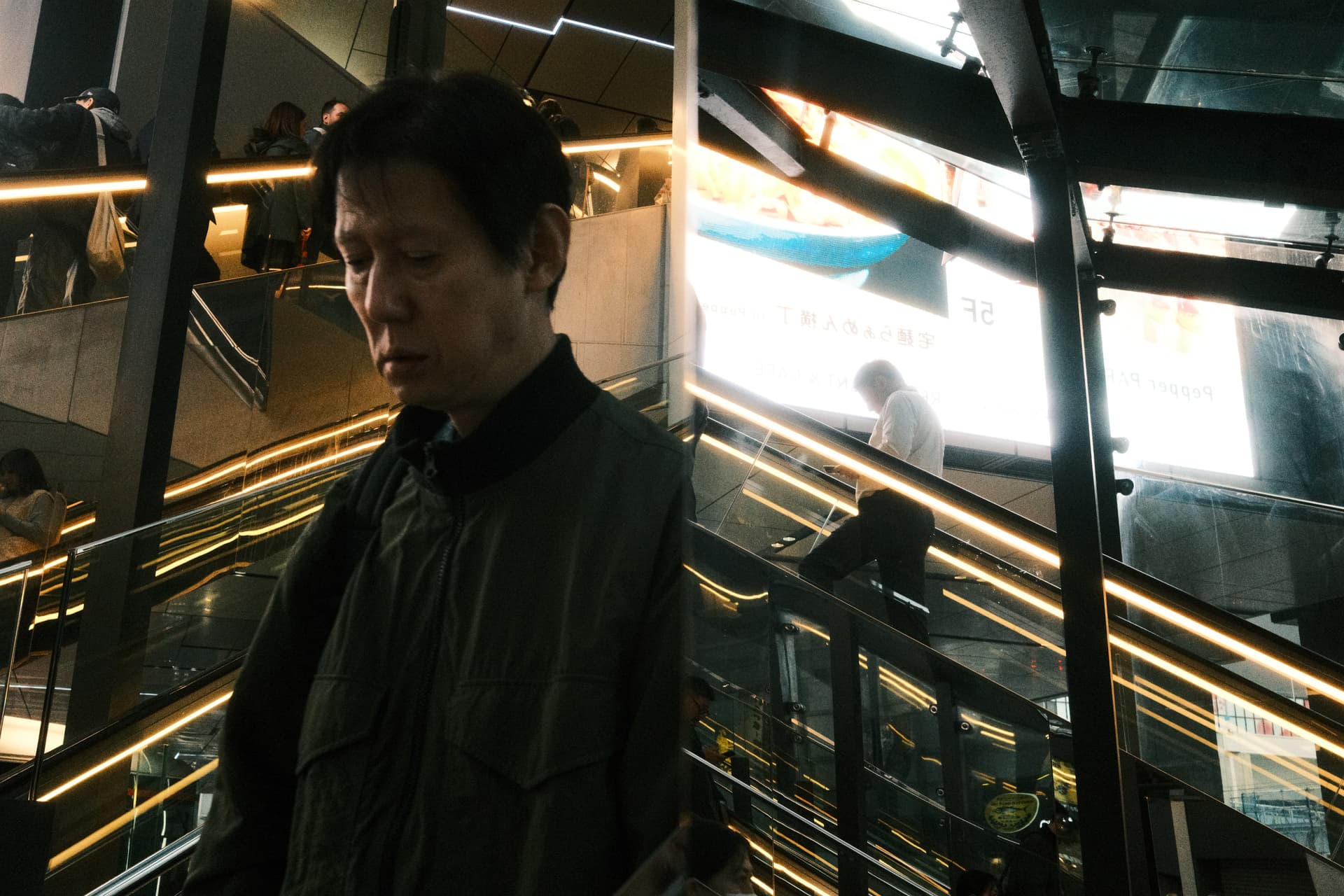 A person stands on a multi-level escalator in a modern Japanese urban space, surrounded by golden-toned LED lighting and glass architectural elements. The moody, low-lit environment suggests an evening scene in a Tokyo transit or commercial building.