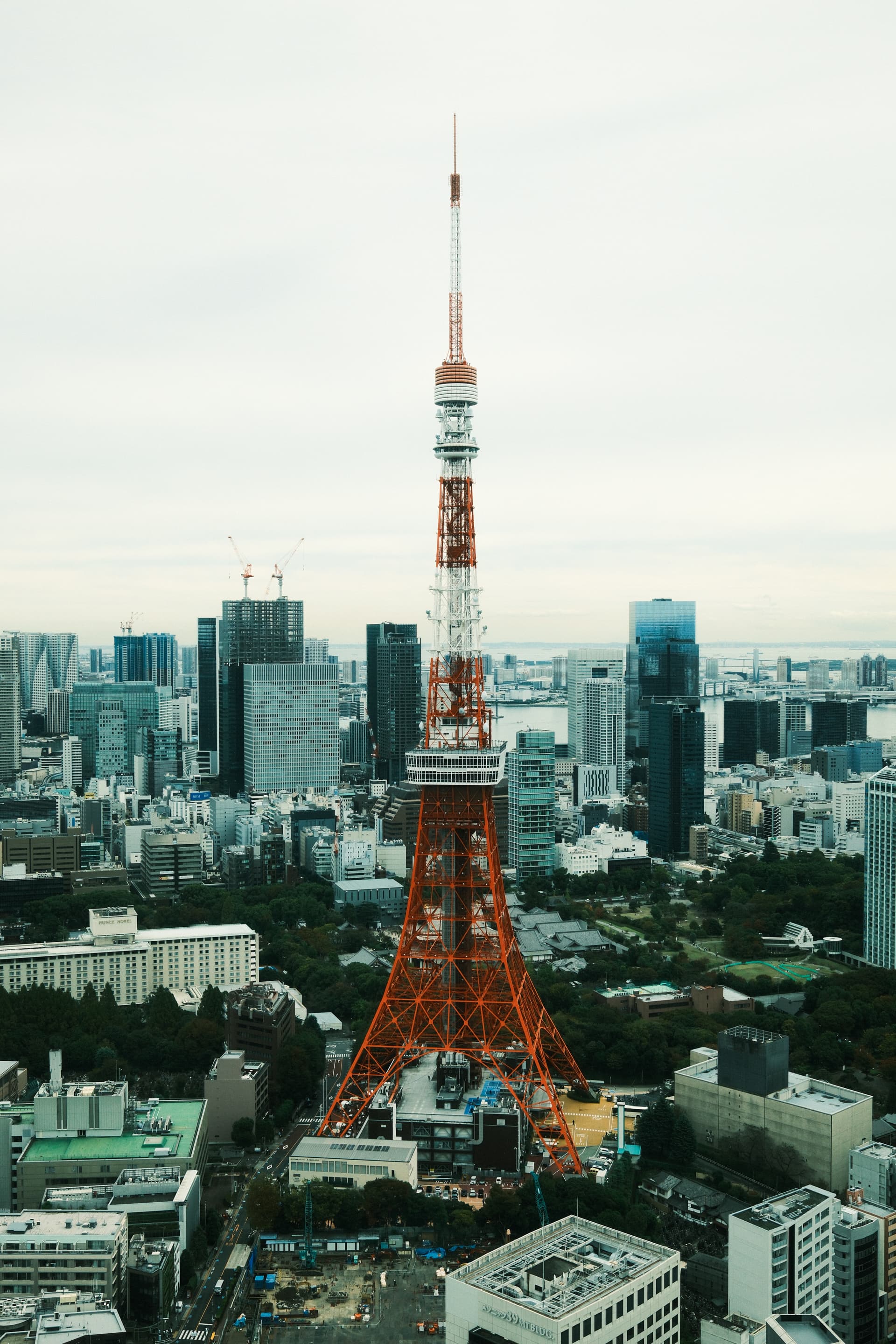 Aerial view of Tokyo Tower, a red and white communications tower resembling the Eiffel Tower, standing prominently among dense urban skyscrapers, green parks, and modern buildings in central Tokyo.