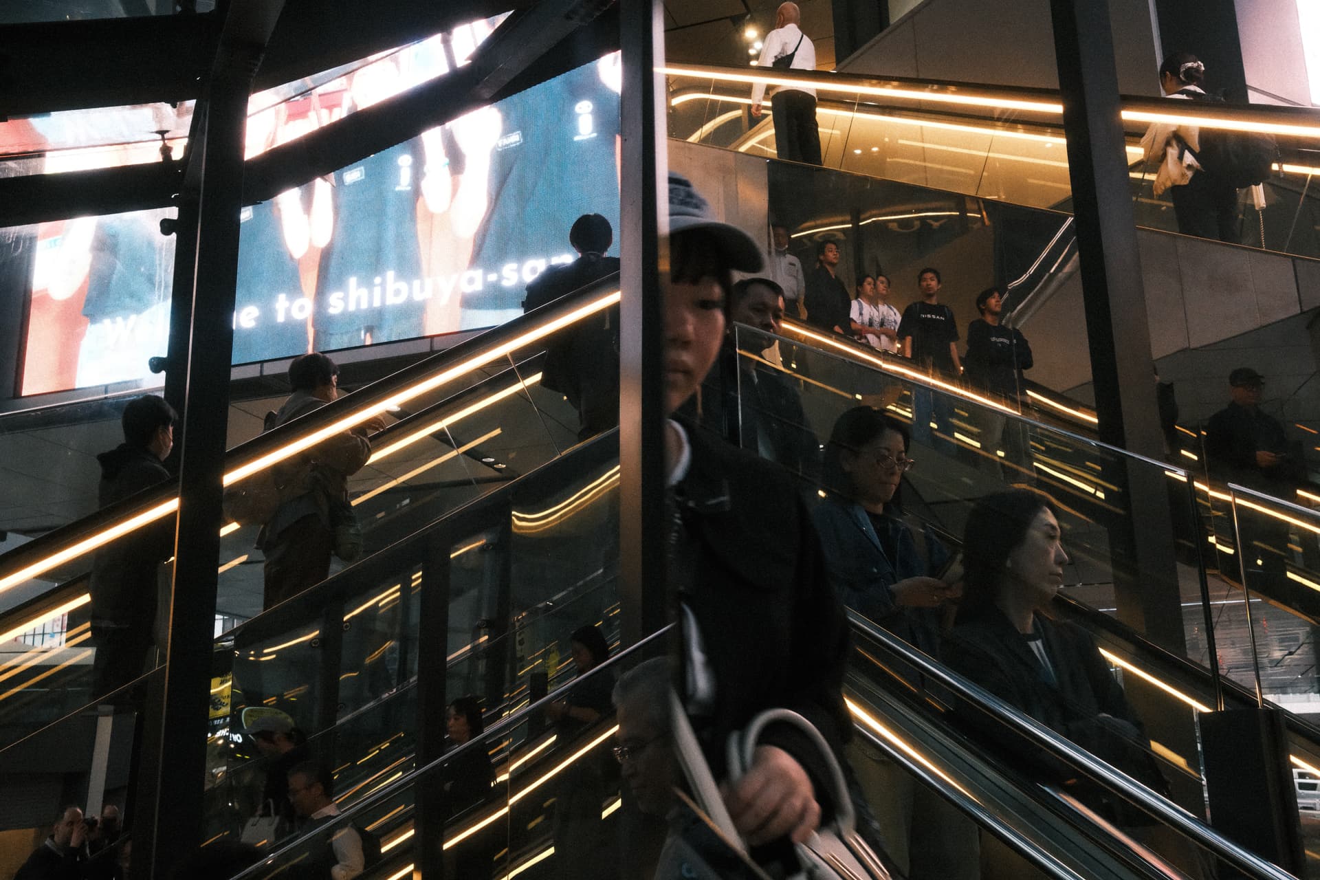 Multi-level escalators with golden lighting in an urban Japanese building, filled with people moving between floors. A large digital screen and reflective glass surfaces create a layered, modern architectural scene typical of Tokyo's contemporary design.
