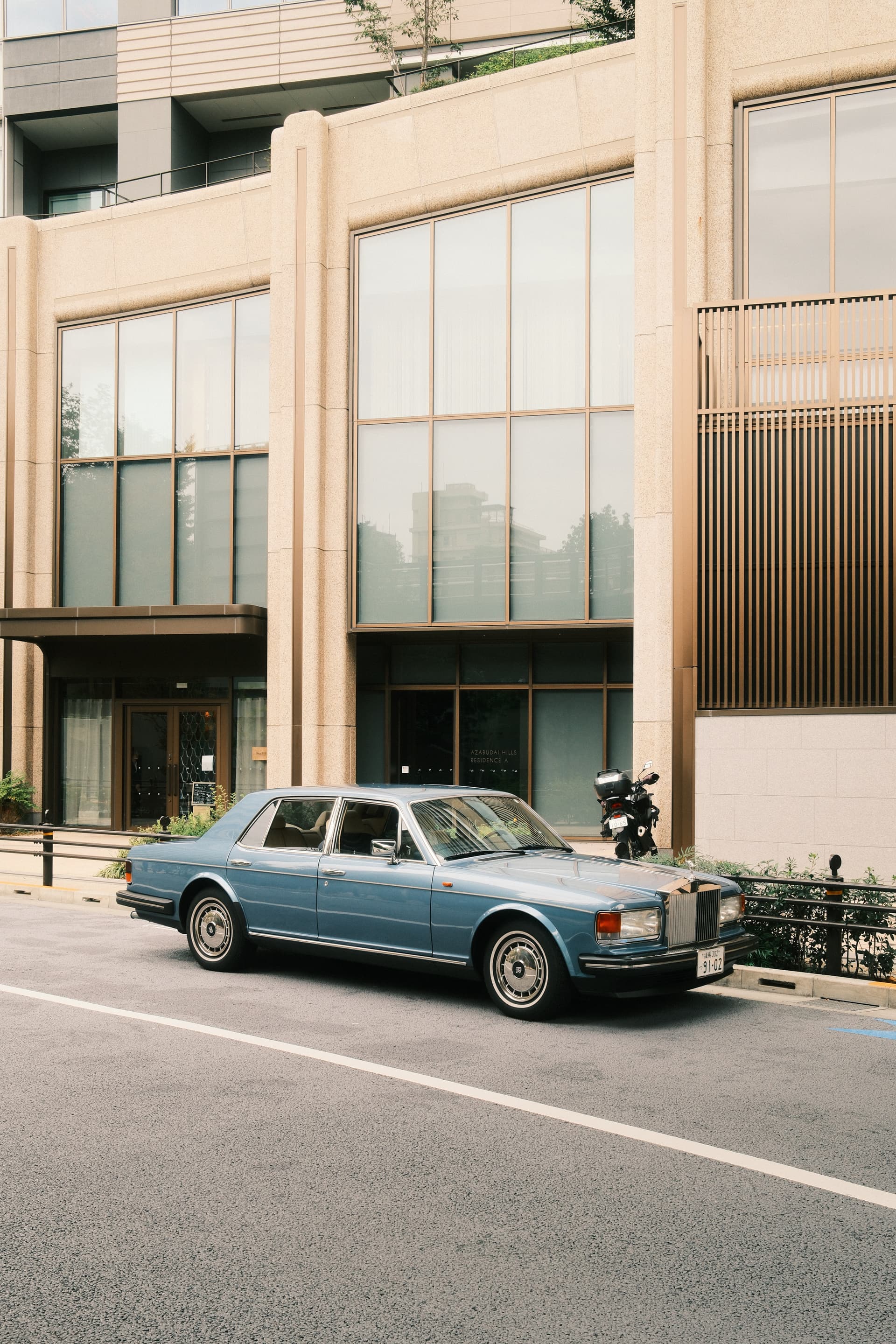A vintage blue-gray Rolls-Royce sedan parked on a quiet Tokyo street, positioned in front of a modern beige building with large glass windows and vertical wooden slat design elements.