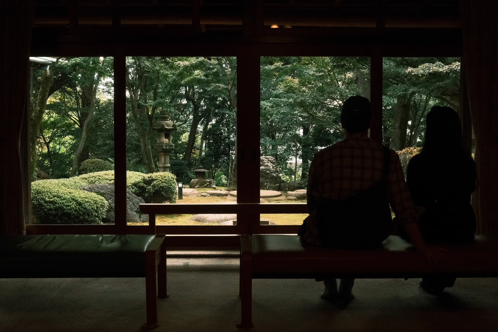 Silhouettes of two people sit in a traditional Japanese interior, looking out large windows onto a lush, green garden with trimmed hedges and a stone lantern, creating a serene, contemplative scene in soft, natural light.